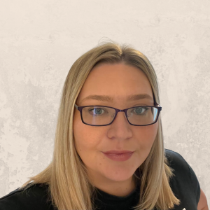 A woman with straight blonde hair, wearing dark-rimmed glasses and a black top, stands in front of a light gray textured background, ready to share top tips for avoiding digital impersonation and cyber fraud scams.